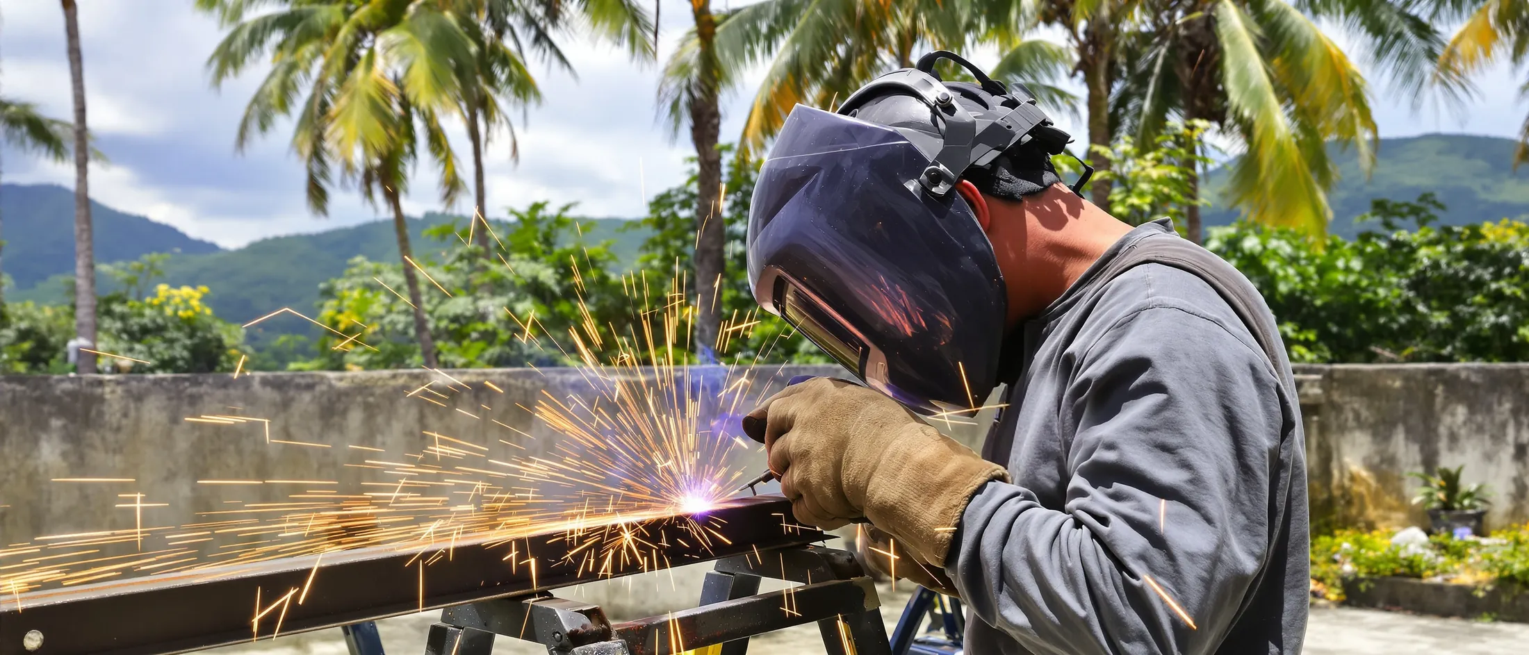 Welder in a welding helmet welding a steel gate frame in a sunny yard, sparks flying, coconut palms and hills behind